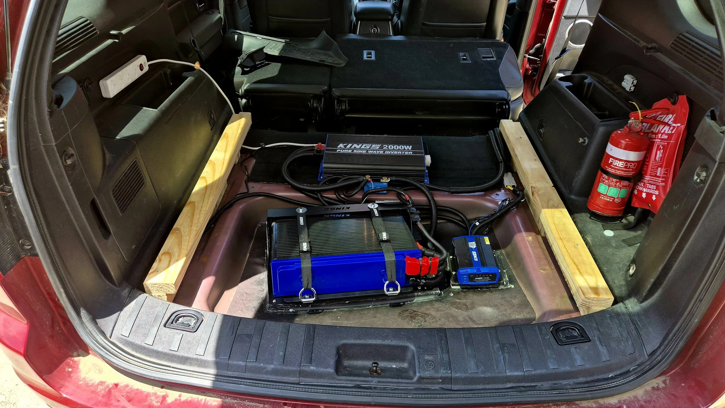 Inside the trunk of a vehicle with battery, inverter, fuses, and fire extinguisher installed, with wooden supports for stability.