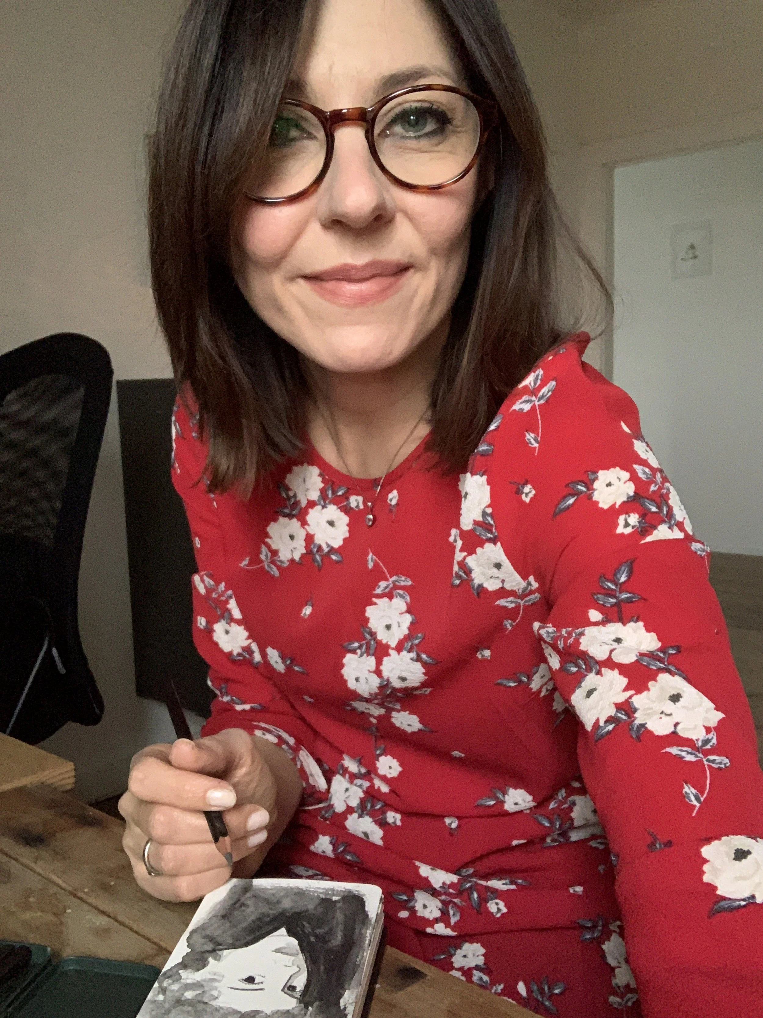 Una donna con occhiali tondi e capelli lunghi scuri, indossa un vestito rosso con fiori bianchi, sorride mentre si prende un selfie in un ambiente interno, seduta a un tavolo.