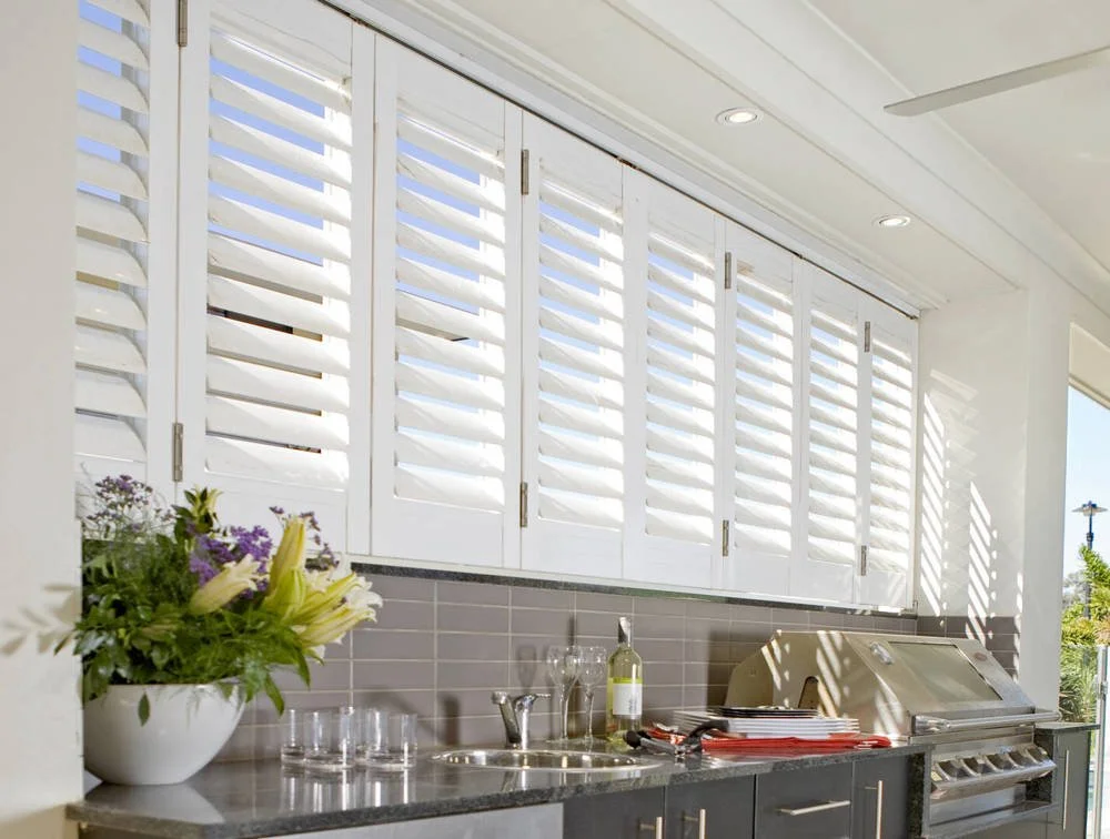 Outdoor kitchen with white shutters, a countertop with a sink, a bottle of wine, glasses, a grill, and a flower vase.