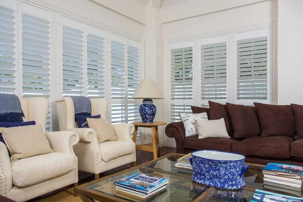 Living room with white armchairs, a brown sofa, a wooden side table, a blue and white vase table lamp, a glass coffee table with magazines, and white plantation shutters on the windows.