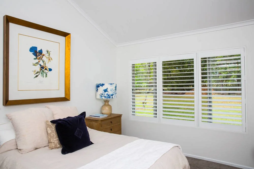 A bedroom with white walls, a bed with pillows, a wooden nightstand with books and a lamp, and large windows with Venetian blinds showing a green yard outside.
