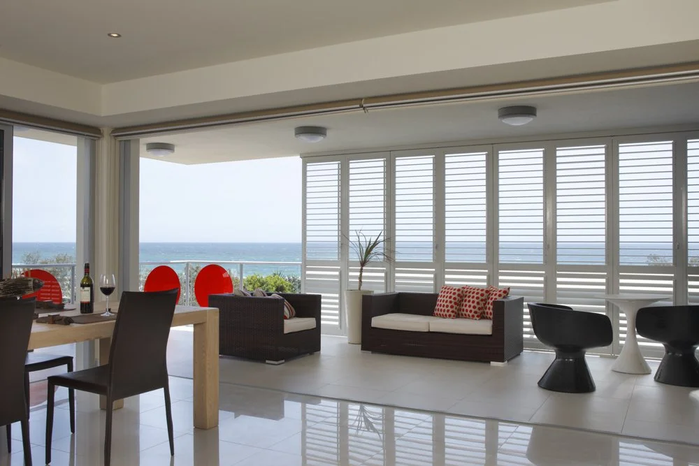 Living room with large windows overlooking the ocean, featuring black couches with red and white cushions, a dining table with wine and glasses, and modern black and white chairs.