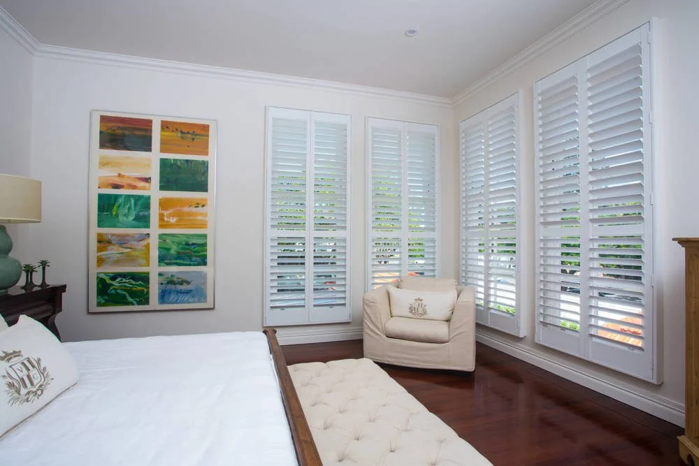 A bedroom with white walls, large windows with white shutters, a wooden bed frame, a white comforter, a matching armchair, a colorful abstract painting, a wooden nightstand, and a wooden dresser.