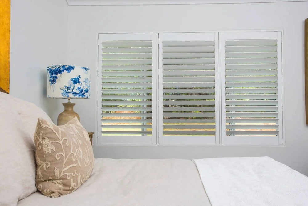 A bedroom window with white plantation shutters, a beige bed with decorative pillows, and a bedside lamp with a blue and white floral lampshade.