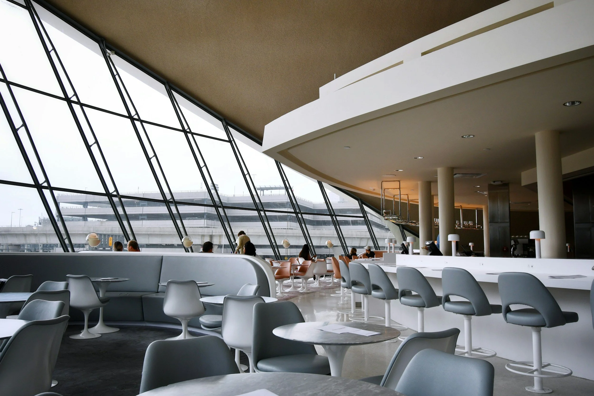 Modern airport lounge with large slanted glass windows, gray seating, white bar with gray chairs, and a few passengers seated and standing.