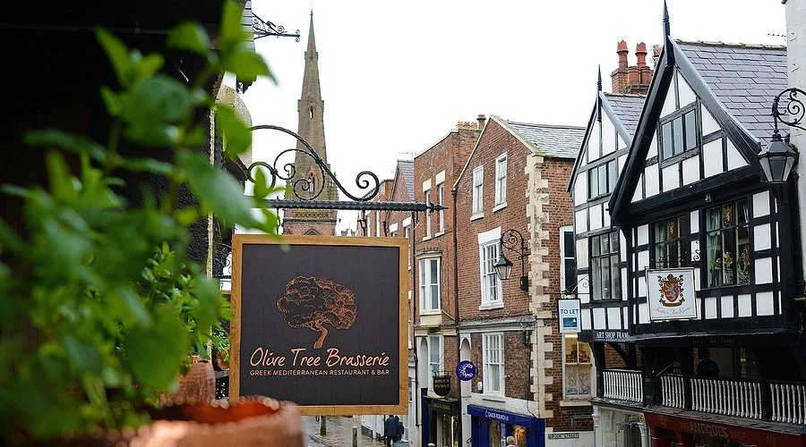 Street view of brick buildings with a black and white Tudor-style building on the right, a sign reading Olive Tree Brasserie, and a church steeple in the background.