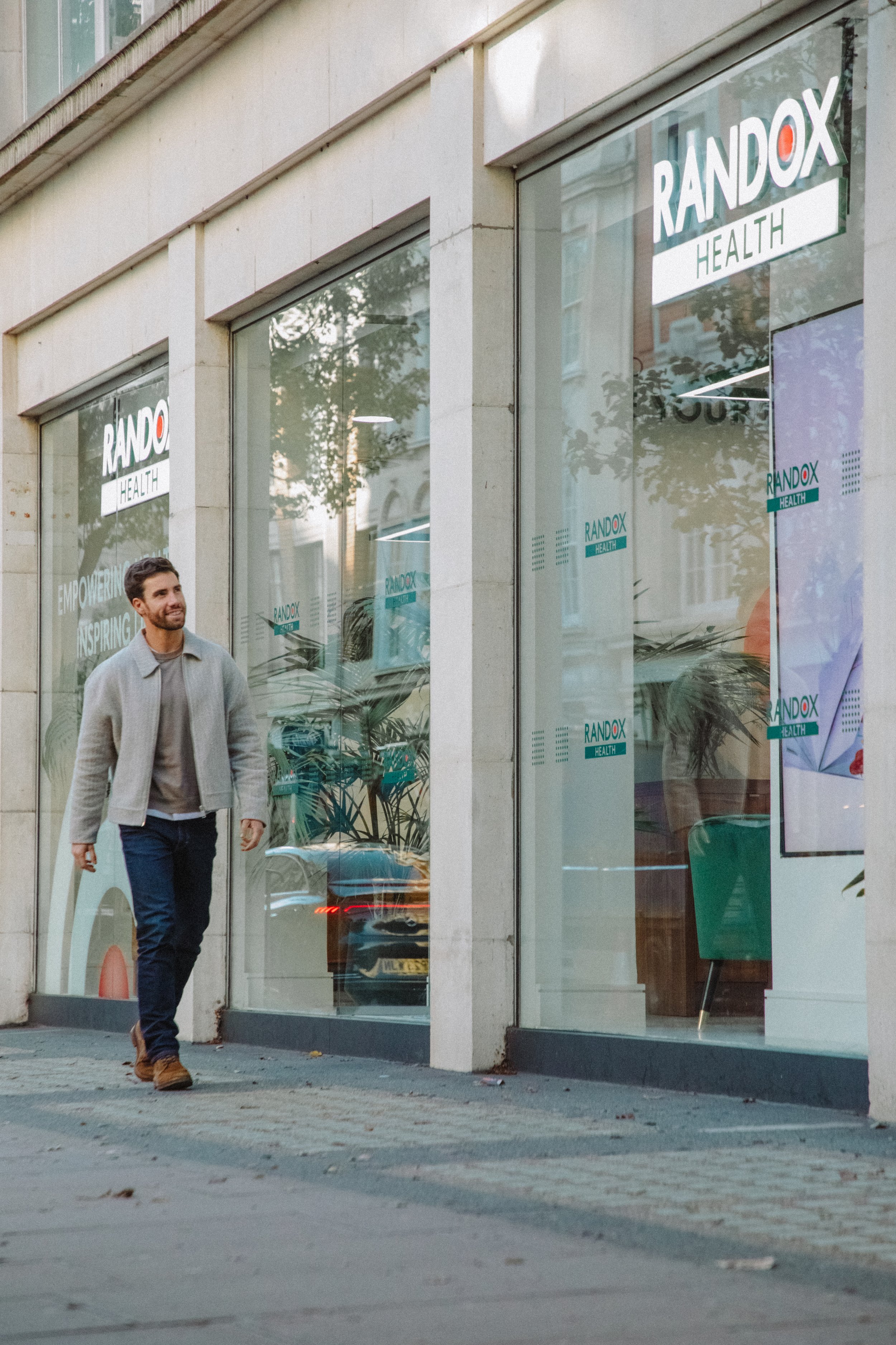 A man walking on the sidewalk in front of a building with large glass windows displaying 'RANDOX HEALTH' signs, trees and reflections visible in the glass.