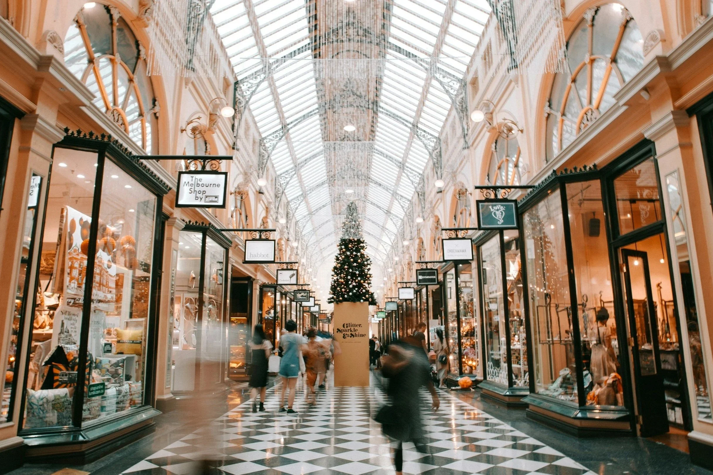 Indoor shopping arcade with a glass ceiling, Christmas decorations, a large Christmas tree in the center, and shops on both sides.