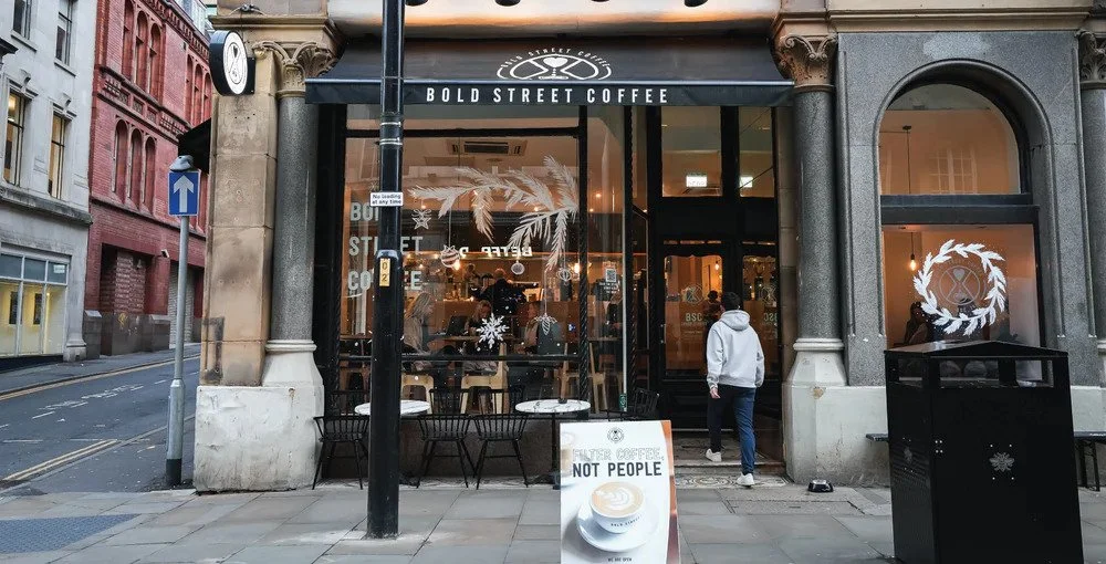 Exterior view of a coffee shop named 'Bold Street Coffee' with large glass windows and a black awning, located on a city street corner, with a person entering the shop, to-go tables outside, and holiday-themed decorations inside.