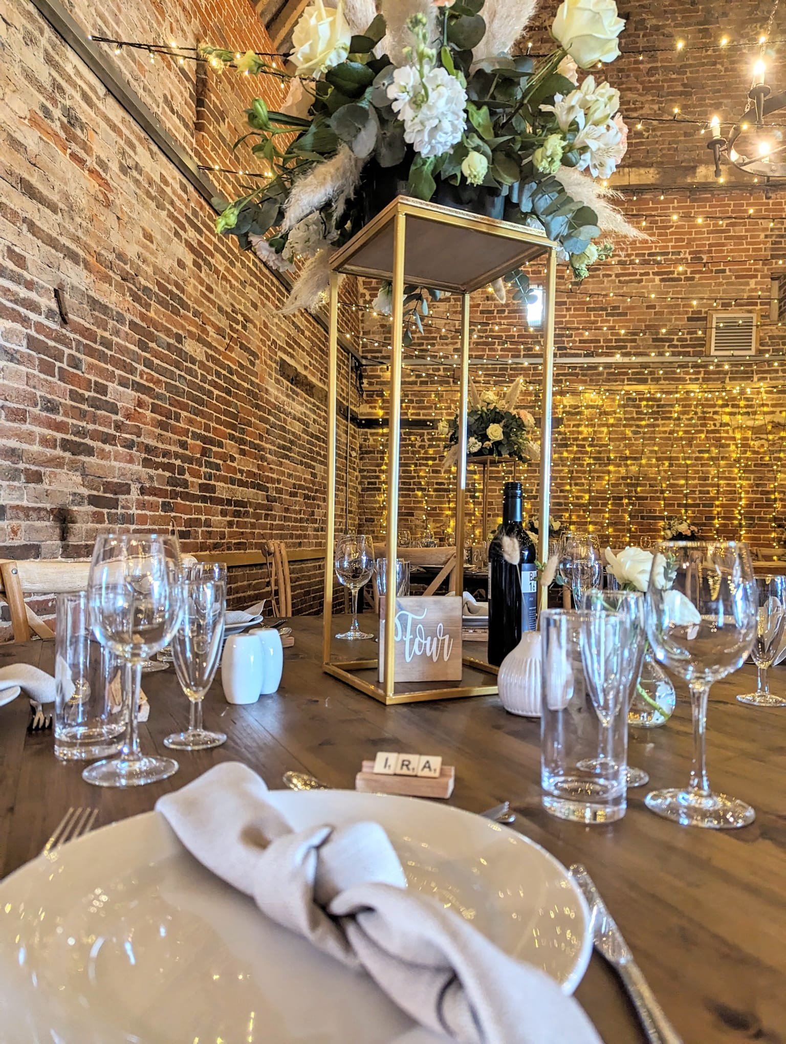 A decorated rustic event table with a floral centerpiece, wine bottles, glasses, and place settings, set against a brick wall with string lights.