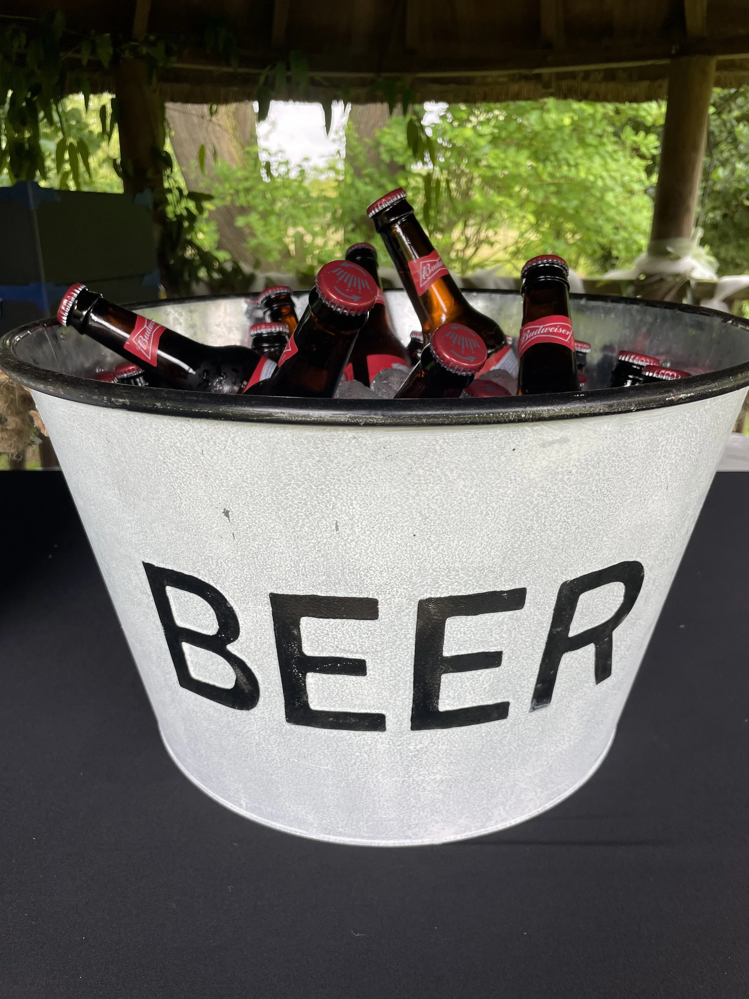 A large frosted ice bucket labeled 'BEER' filled with ice and several bottles of Budweiser beer, set on a black surface with a green outdoor background.
