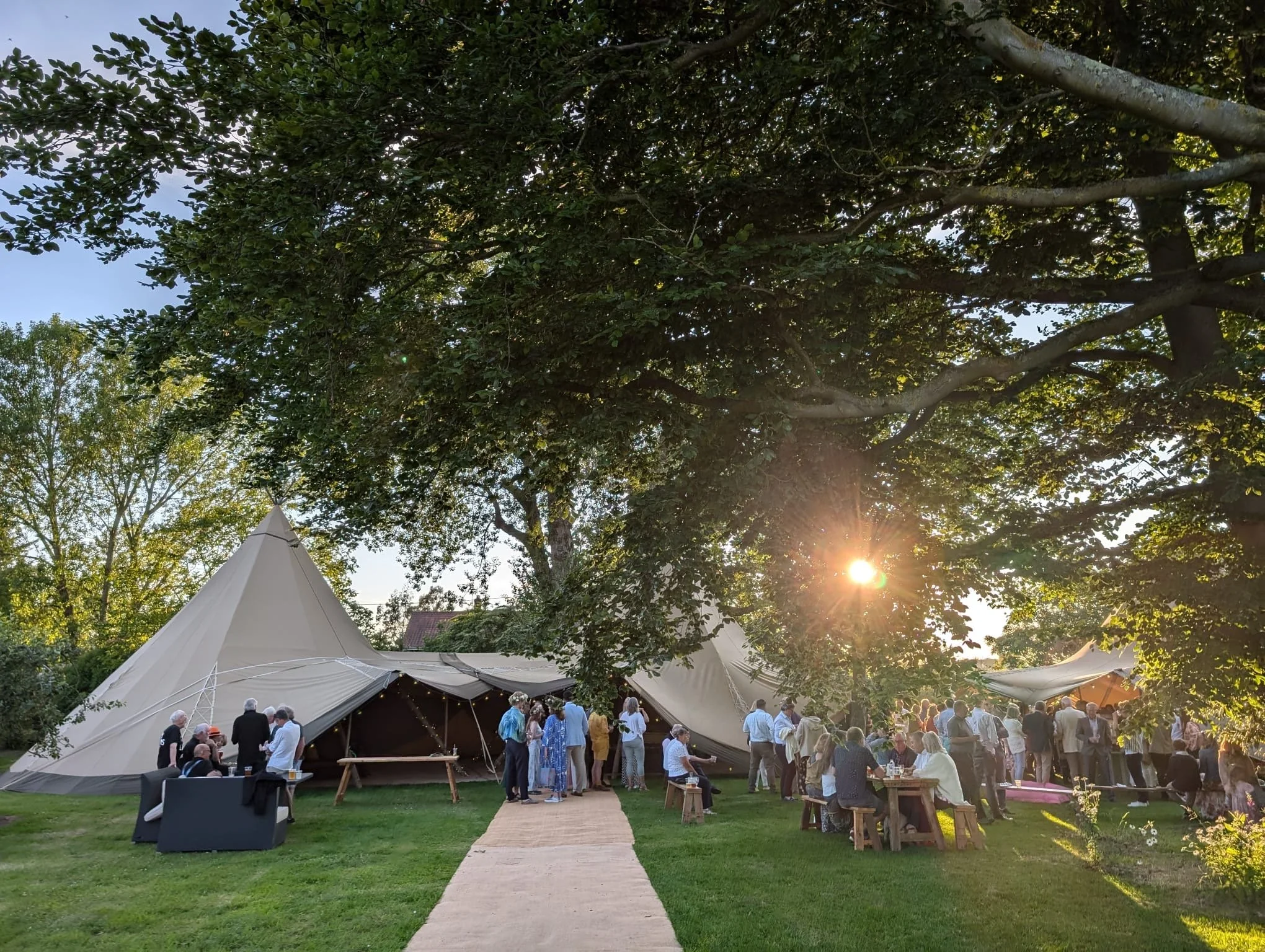 An outdoor gathering with people socializing near large white tents, sitting at picnic tables, under trees with the setting sun shining through the branches.
