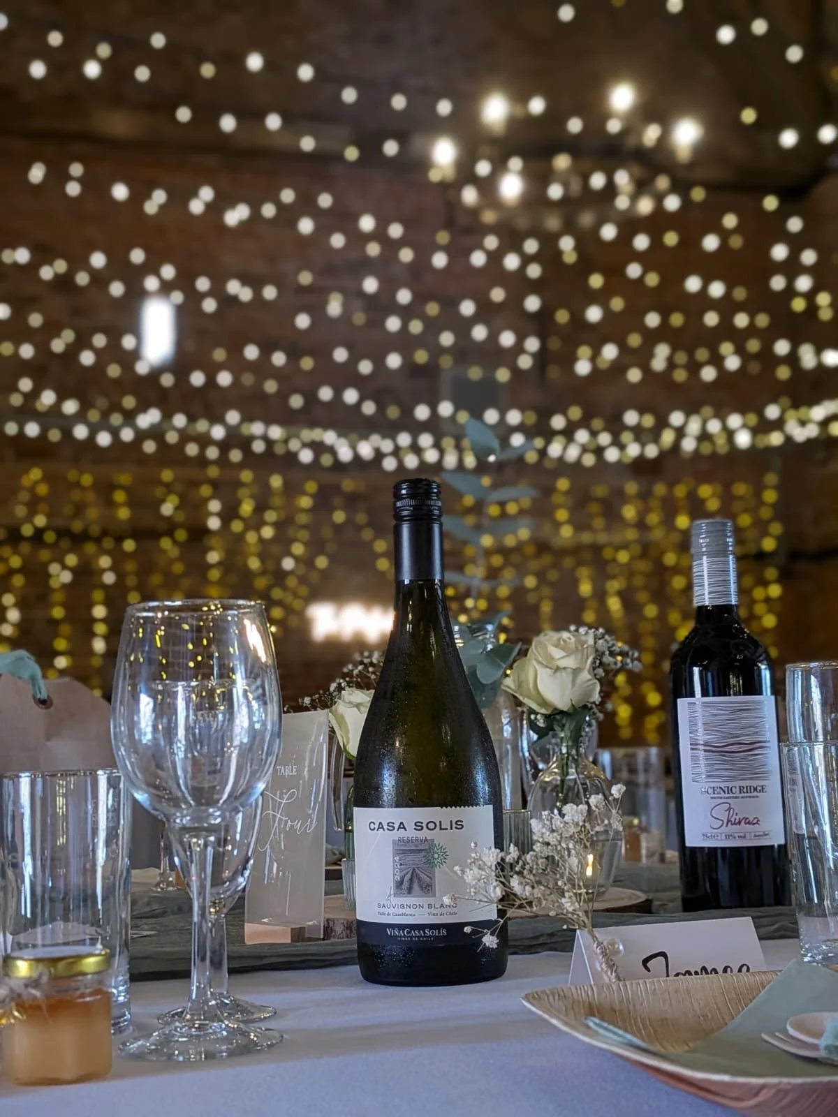 Table setting with wine bottles, glasses, flowers, and a candle, with a background of blurred string lights and a rustic ceiling.