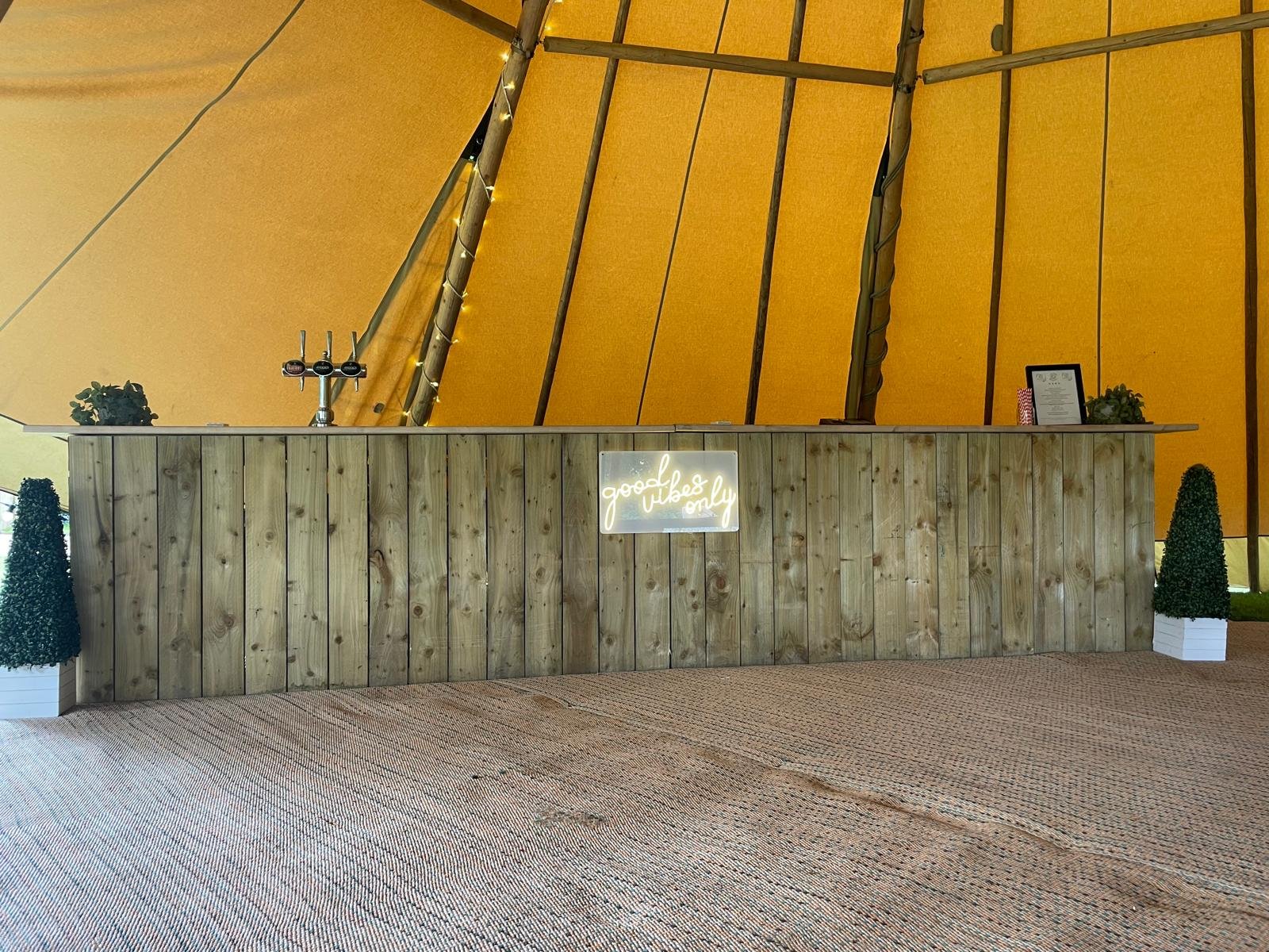 Wooden bar counter with a neon sign that says "good vibes only," four beer taps, framed certificate, plants, and small decorative items on the counter, with an orange canopy overhead.