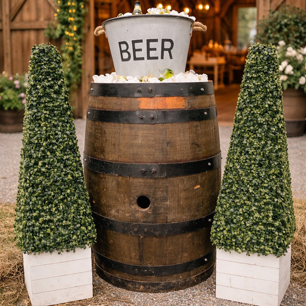 A wooden barrel with black metal bands, topped with a metal bucket labeled 'BEER' filled with ice, surrounded by two tall, trimmed green topiary trees in white square planters, set outdoors with a wooden fence and string lights in the background.