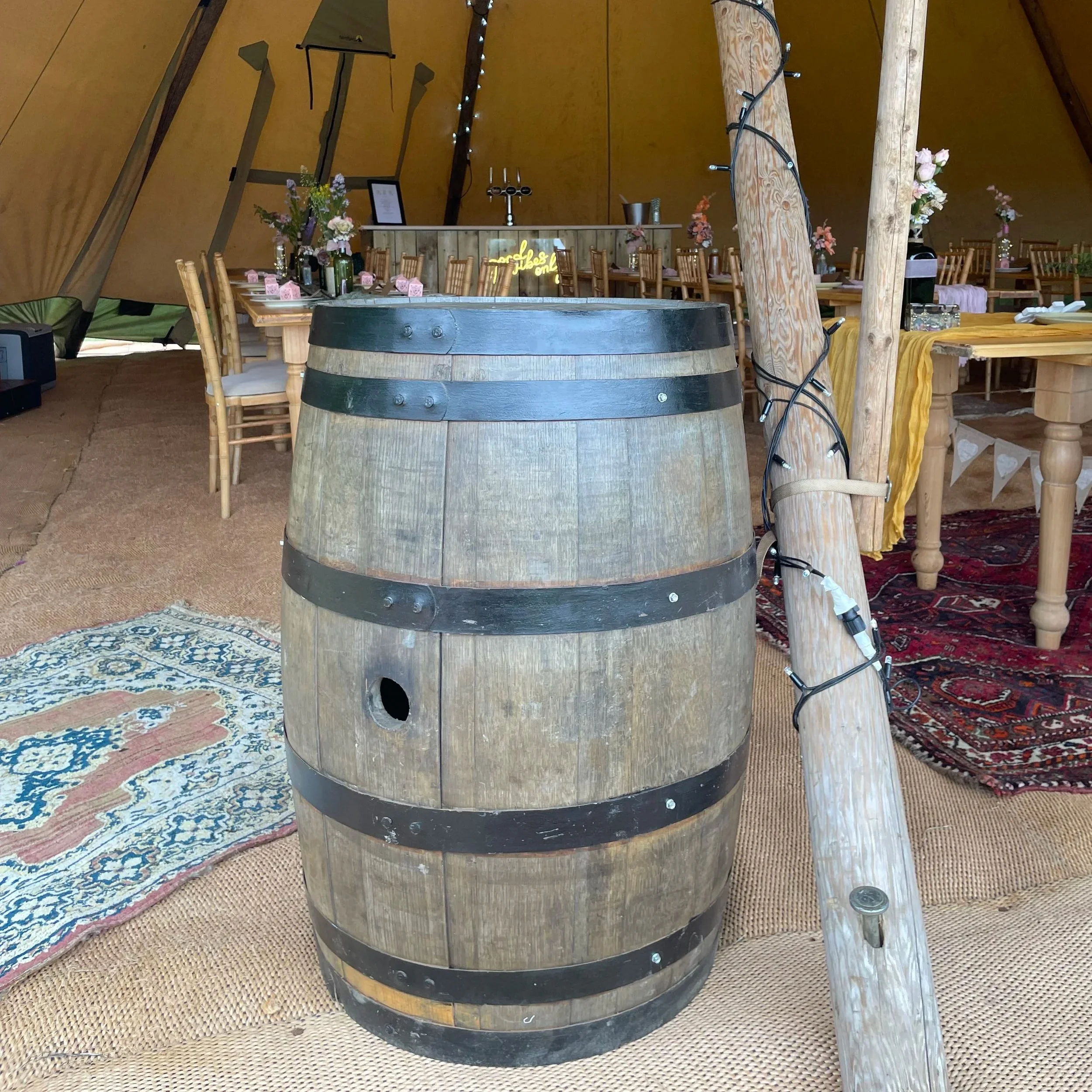 A wooden barrel with metal bands in a decorated event space, with a tent interior showcasing tables, chairs, floral centerpieces, and string lights.