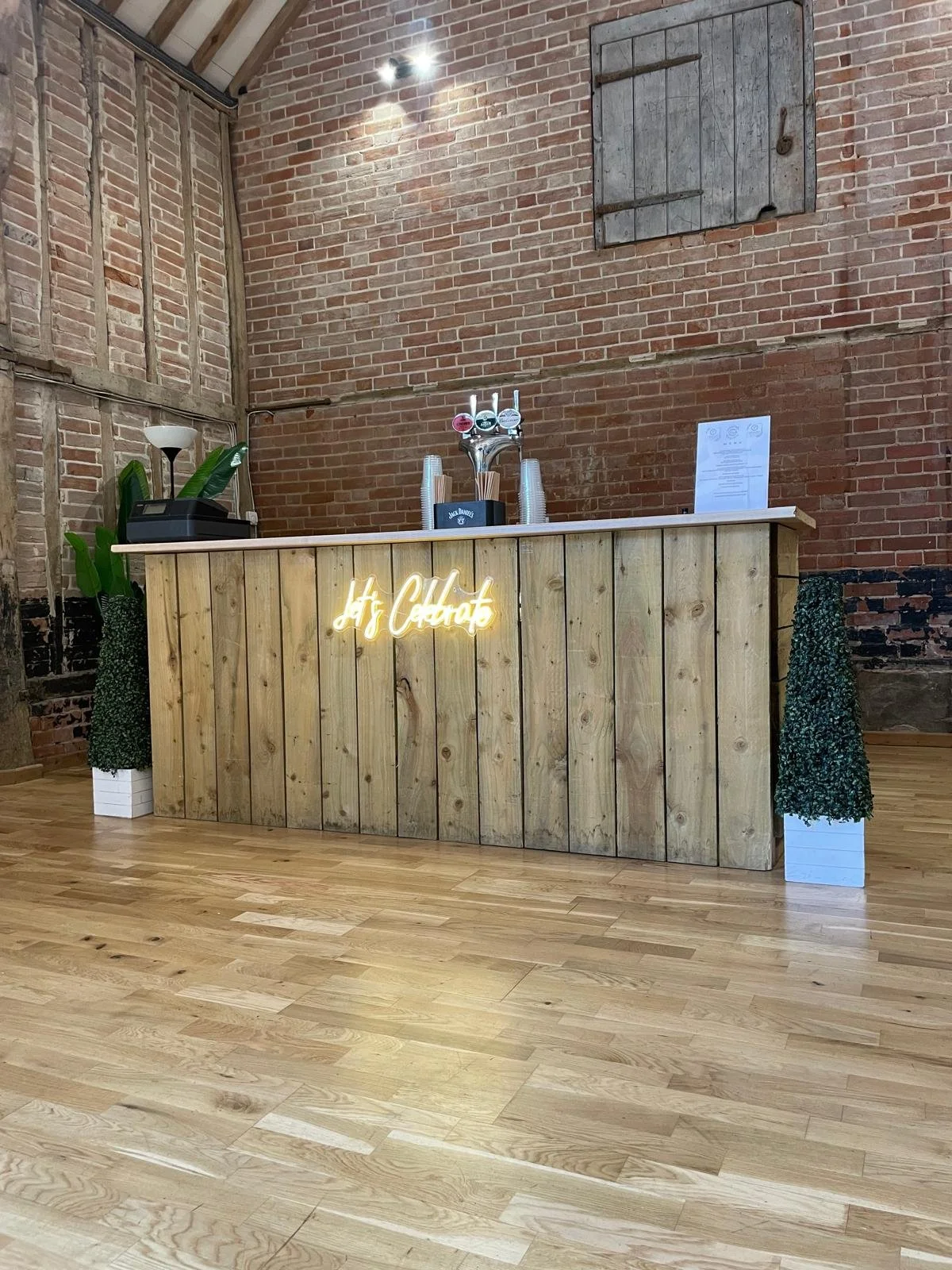 A wooden bar with a neon sign that reads 'Hyg Ciders'. There are two planters with green plants on either side of the bar, metal cups, and a beer tap with three handles on top of the bar. The background features a brick wall with a small shutter and a paper sign.