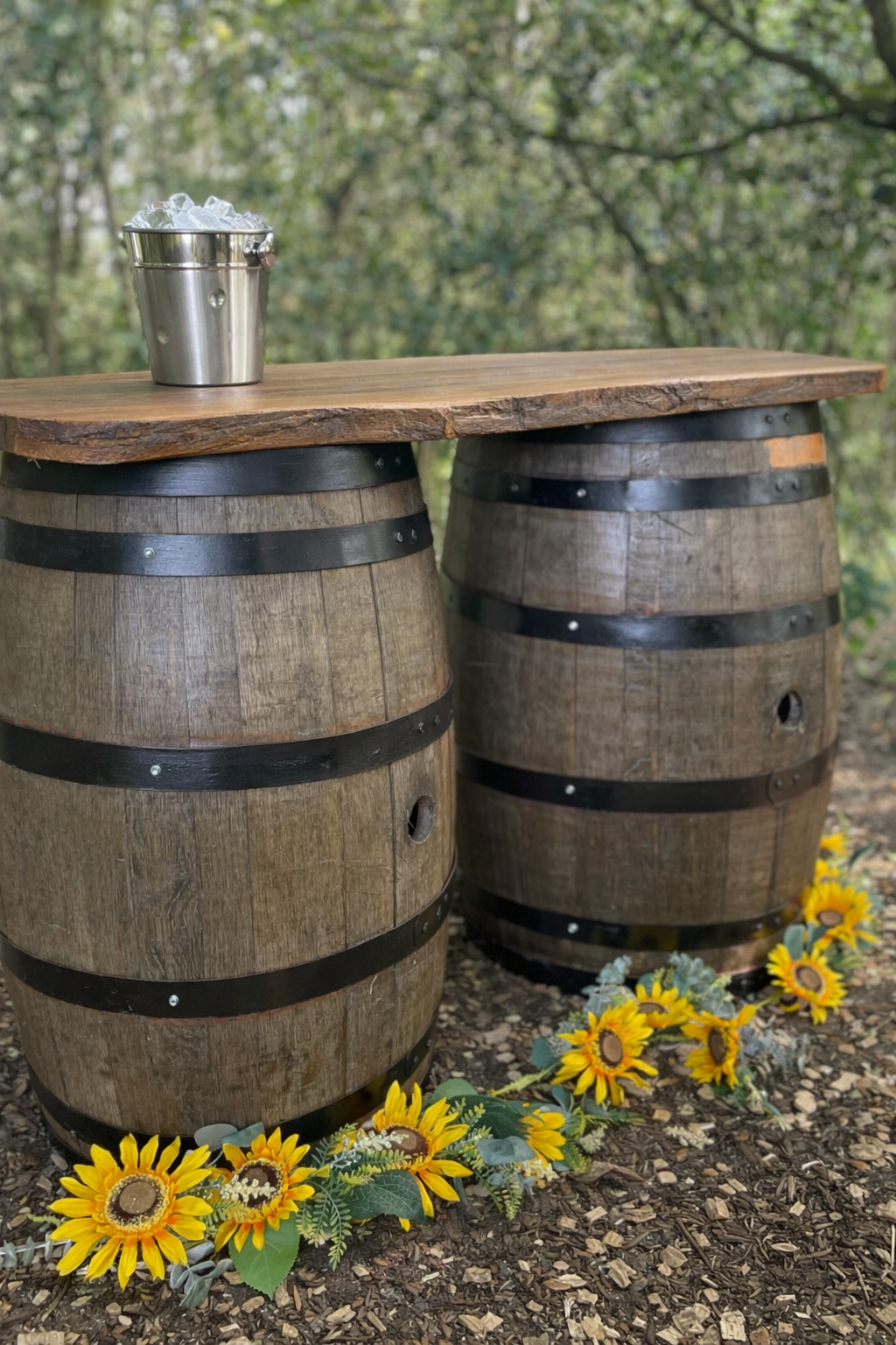 A rustic outdoor table made from two wooden barrels with a wooden top, surrounded by yellow sunflowers on the ground, set in a wooded area.