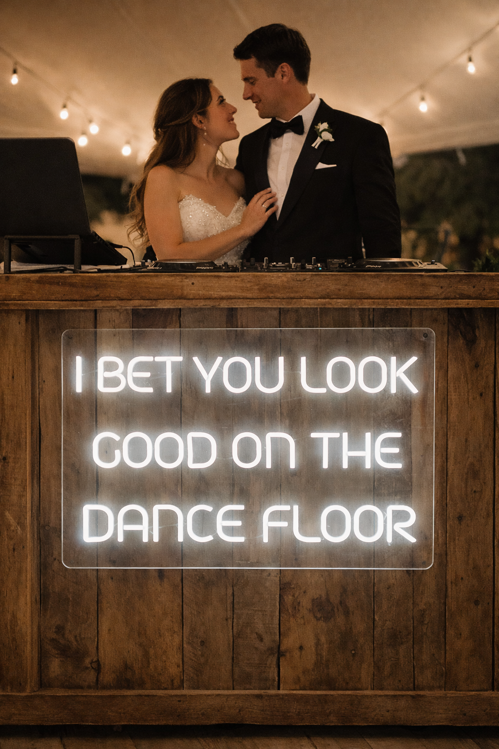 A bride and groom in wedding attire are dancing close together behind a DJ booth with a neon sign that reads, "I bet you look good on the dance floor," in a warmly lit venue.
