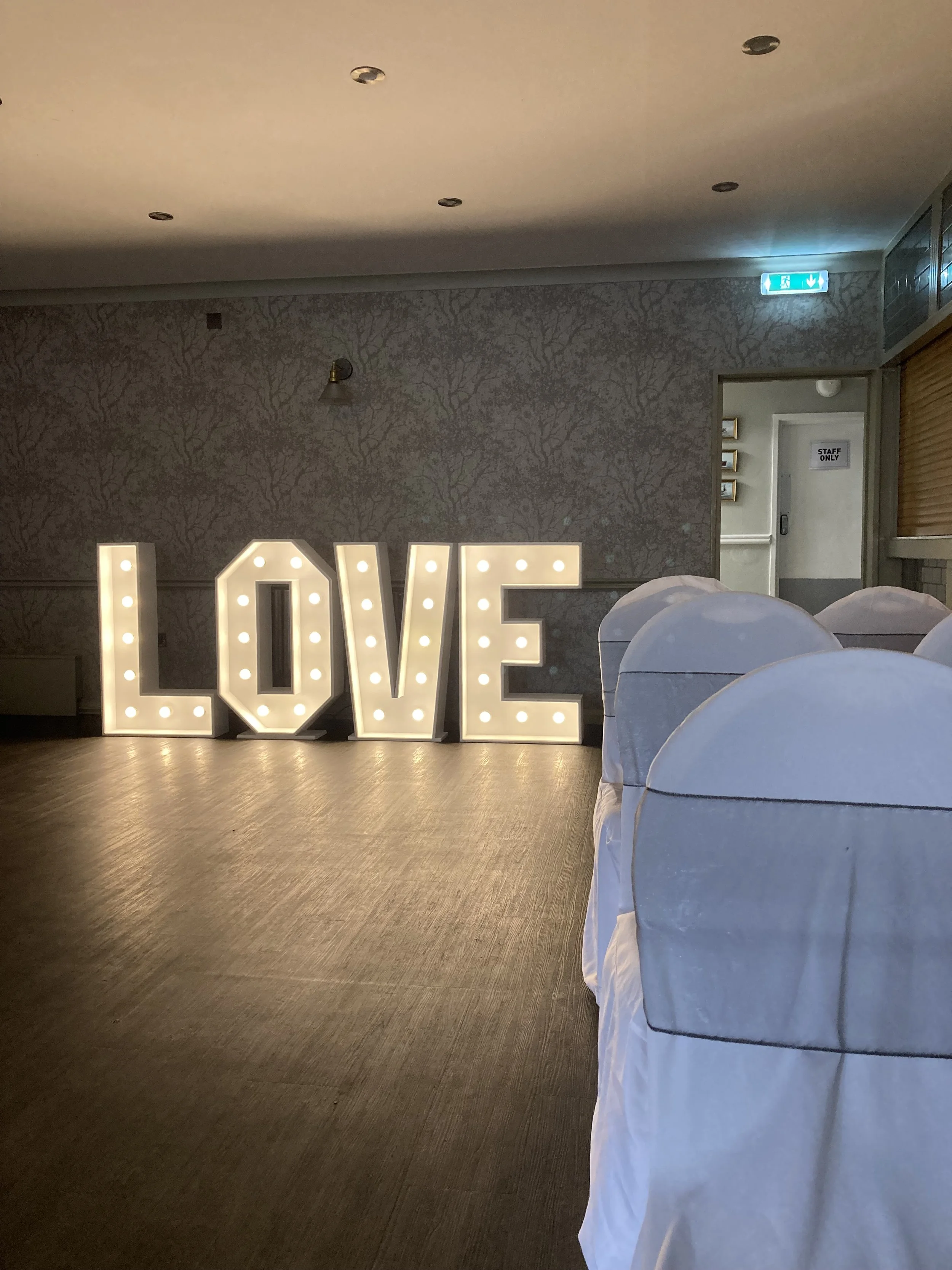 Illuminated marquee sign spelling out 'LOVE' on a wooden floor at an event with white chairs covered in white fabric and a patterned wall in the background.