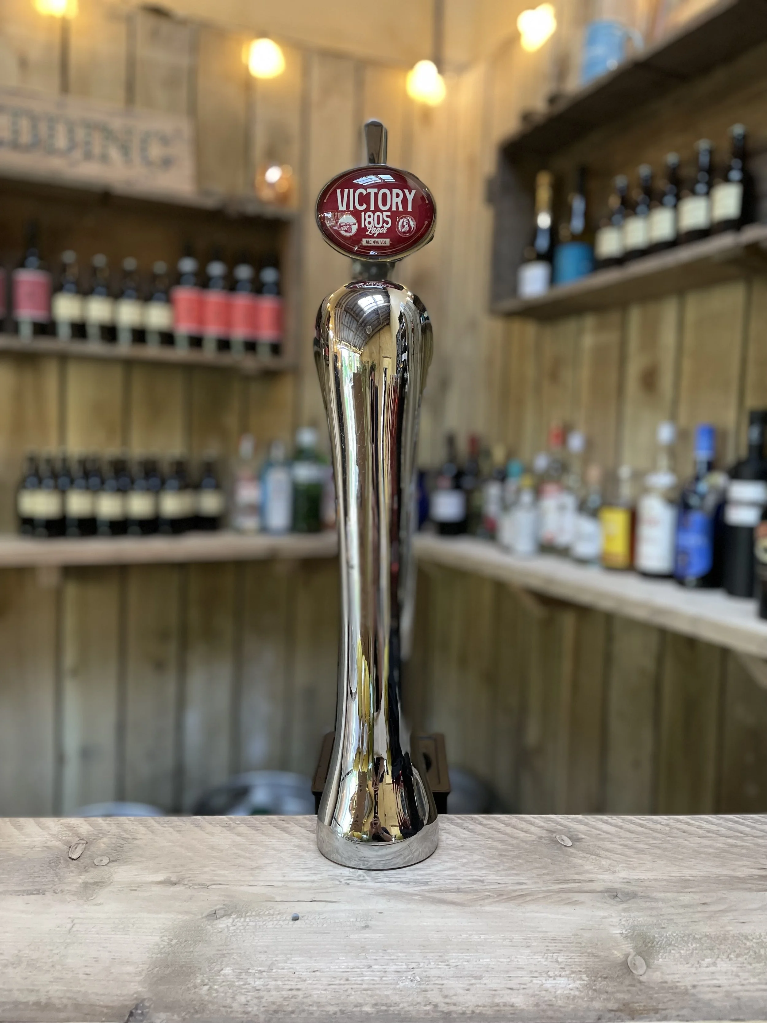 A metal beer tap with a red label reading 'Victory 1805 Lager' on top, located on a wooden bar counter, with shelves of bottles and a wooden wall in the background.