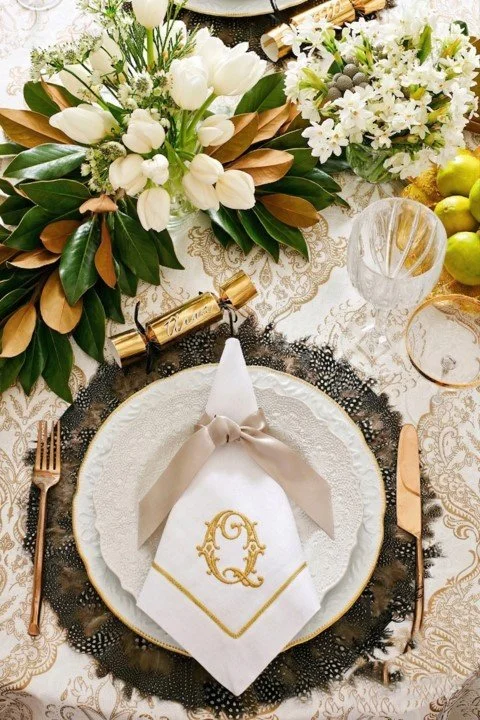 Elegant table setting with a white napkin embroidered with a gold letter, set on a white plate with a black lace charger, surrounded by gold cutlery, floral arrangements with white flowers and greenery, and decorative items including candles and green apples.