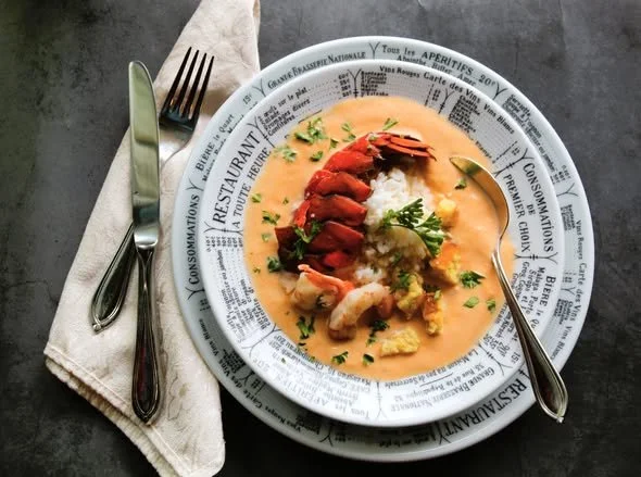 Lobster dish with corn and herbs in a creamy sauce, served on a white plate with a newspaper print rim, accompanied by a fork and knife on a napkin.