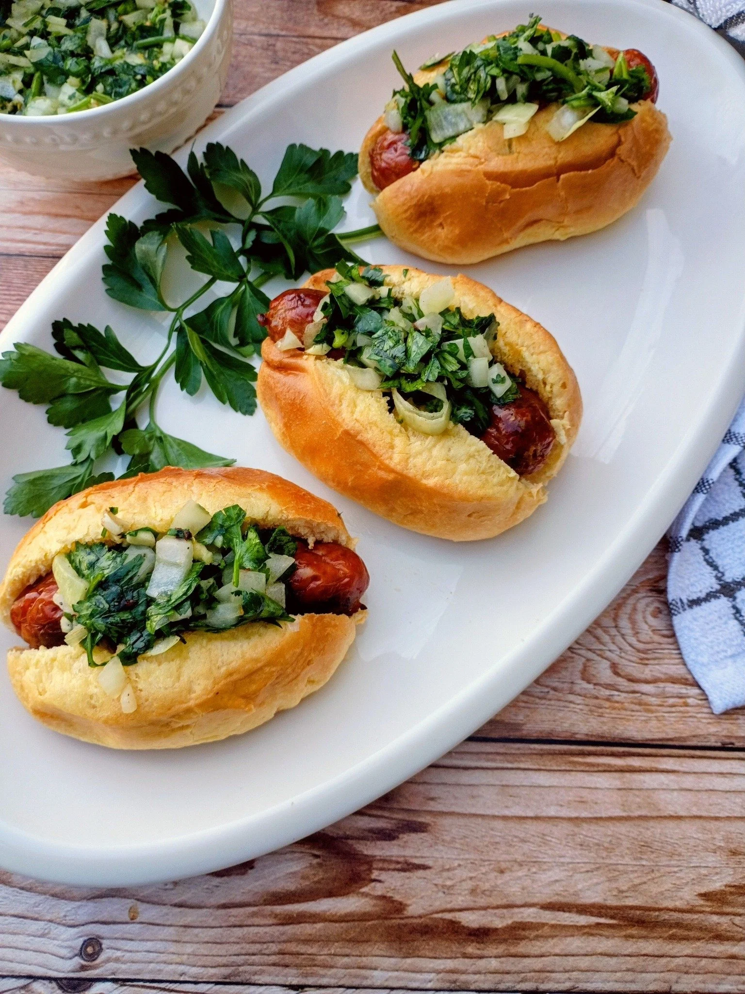 Three hot dogs topped with chopped onions and greens on a white platter, garnished with parsley, on a wooden table.