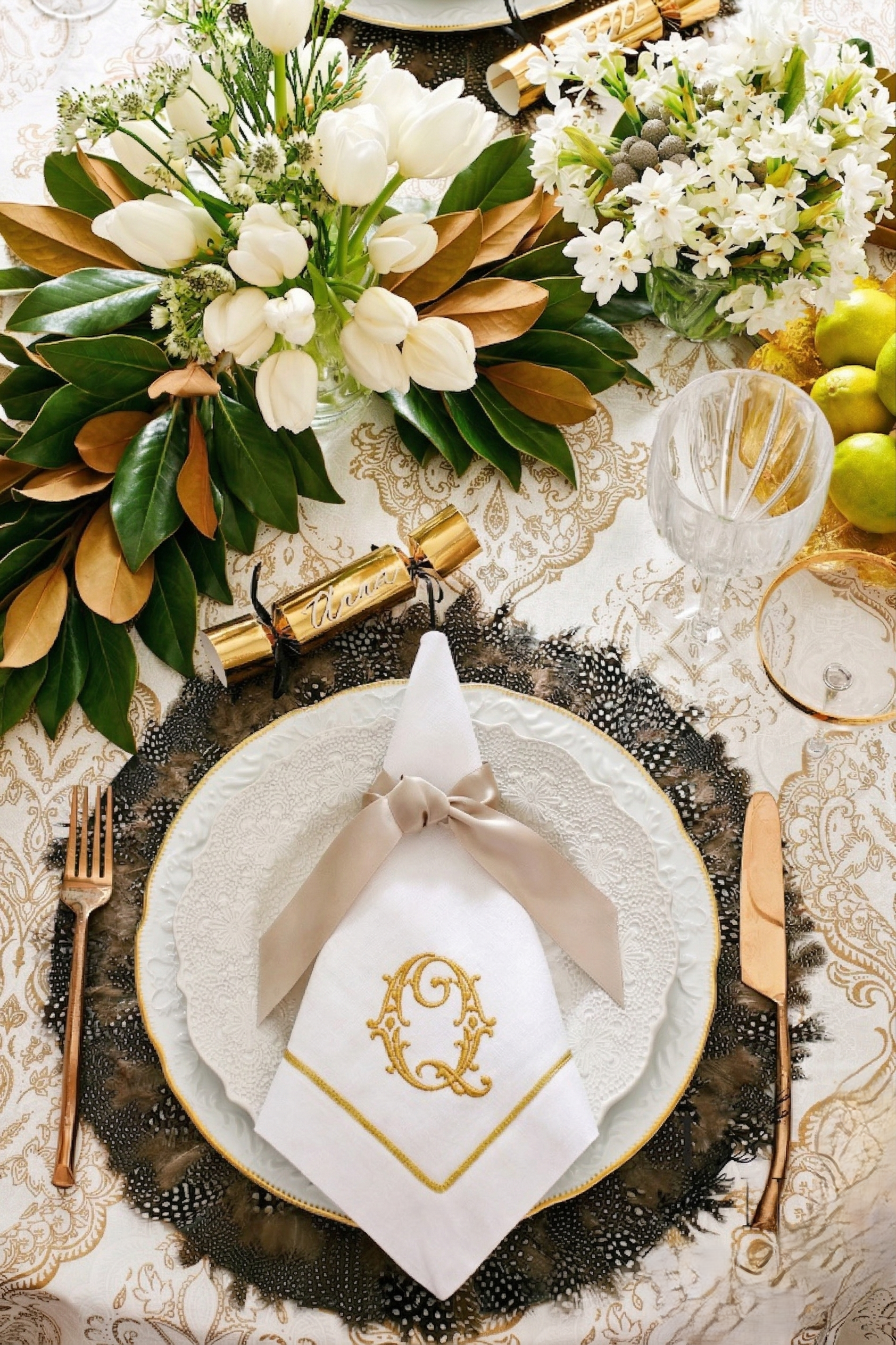 Elegant table setting with white embroidered napkin and gold initials, floral centerpiece with white tulips and greenery, gold cutlery, crystal glassware, and a decoration with a gold train, on a gold and white patterned tablecloth.