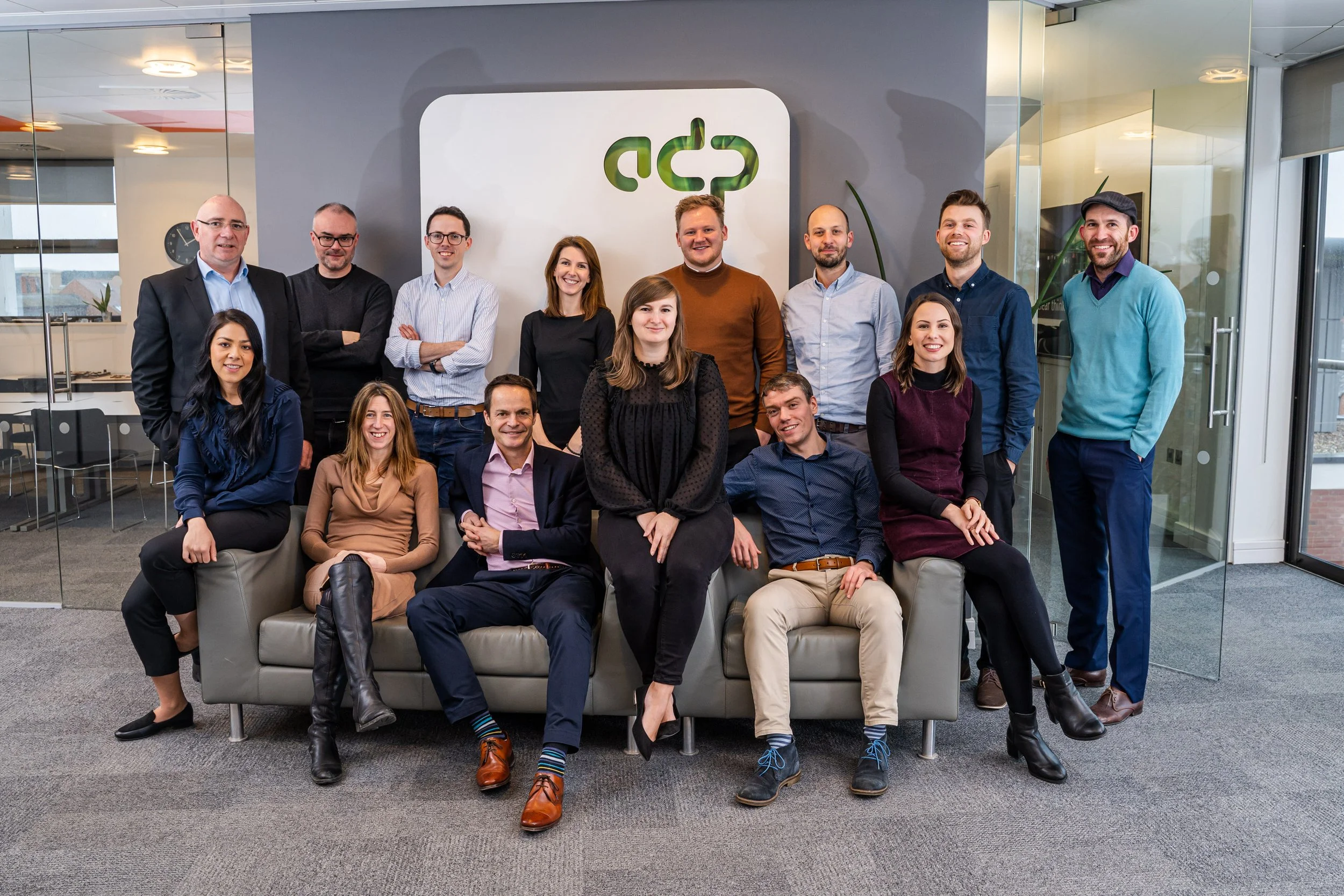 Group of 14 diverse professionals in business casual attire, posing for a team photo in an office with a logo on the wall.