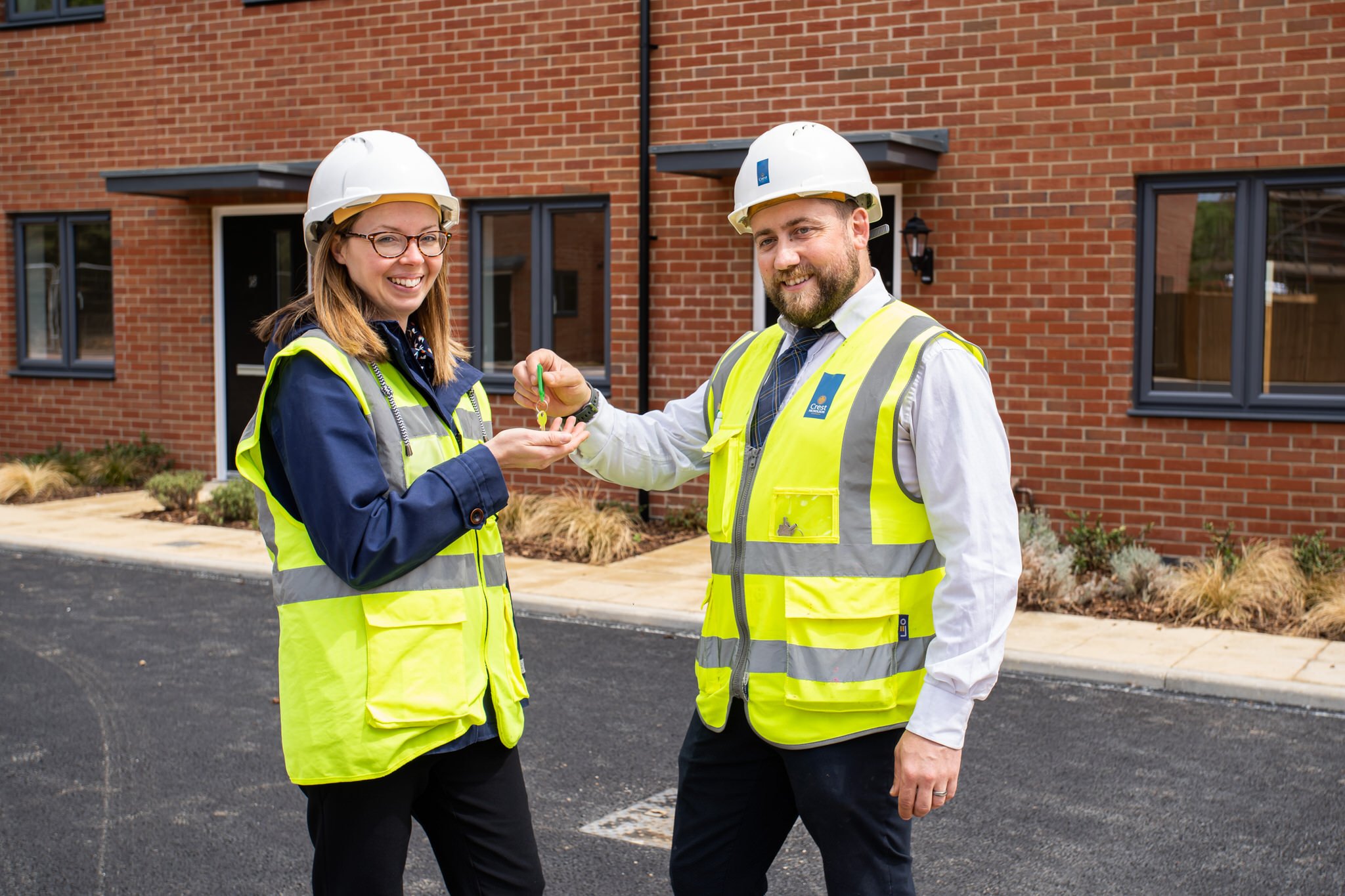 Two construction operatives, a woman and a man, smiling outdoors in front of a brick building. The woman is receiving house keys from the man. Both are wearing white hard hats and yellow safety vests.
