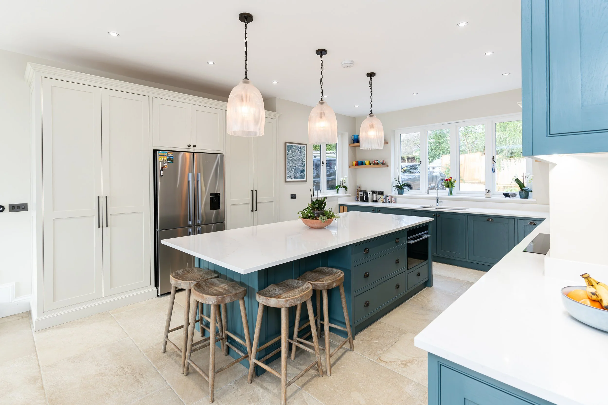 Modern kitchen with white countertops, teal cabinetry, an island with four wooden stools, built-in stainless steel refrigerator, and large windows bringing in natural light.