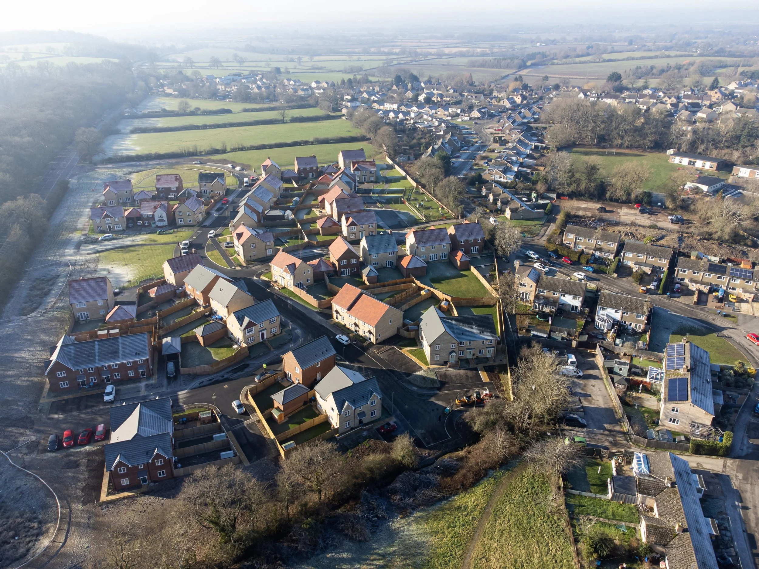 Aerial view of a residential neighborhood with numerous houses, green lawns, roads, and some trees, set in a rural area with open fields in the background.