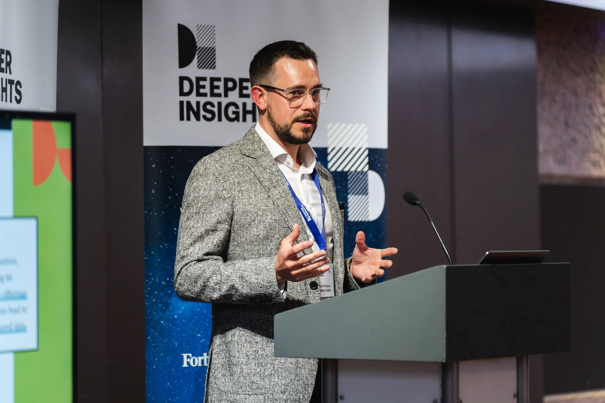 Man in gray blazer and glasses giving a presentation at a conference with a podium and a Deep Insight banner in the background.