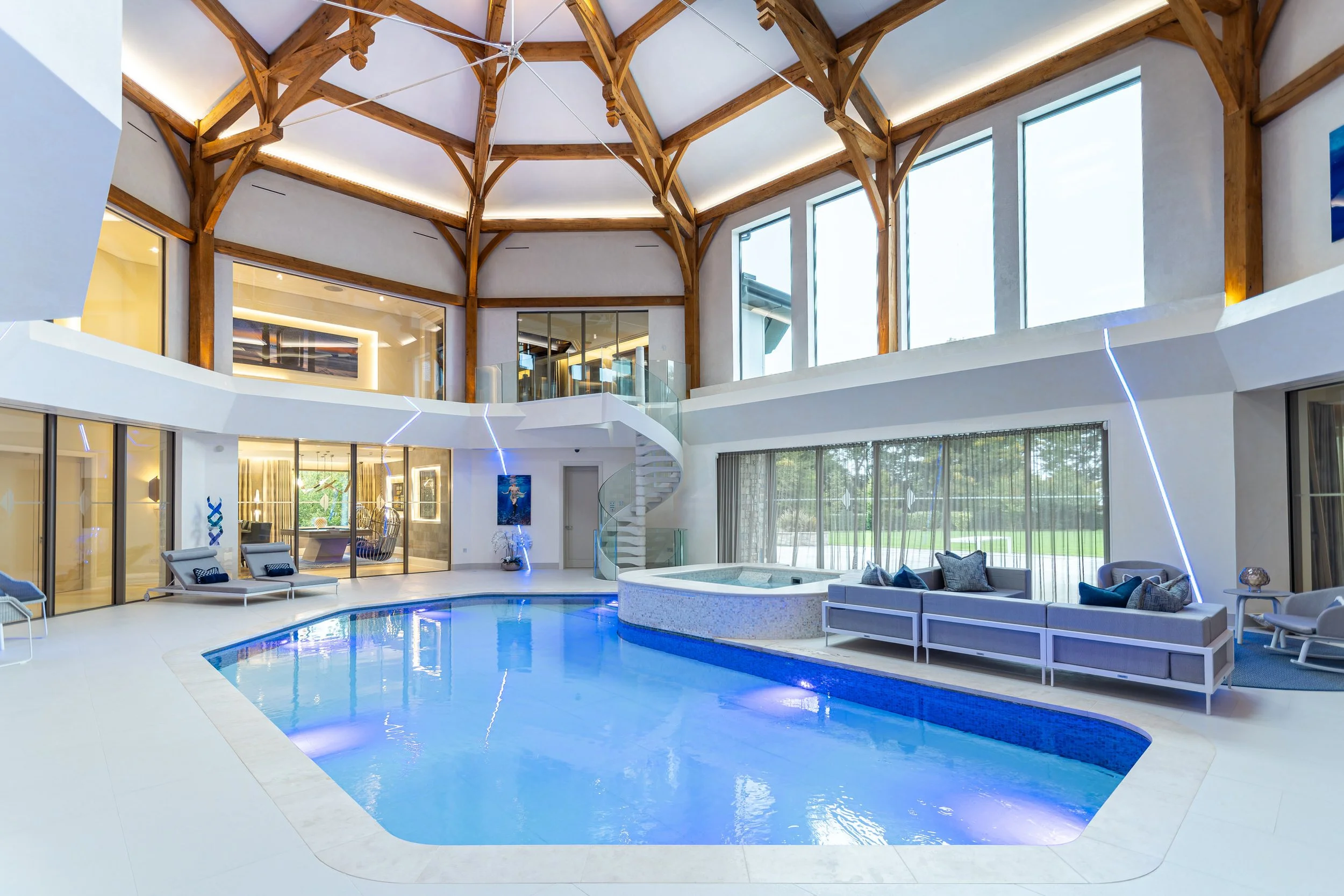 Indoor swimming pool area with modern design, featuring a curved pool, large windows, wooden ceiling beams, and seating area with couches and lounge chairs.