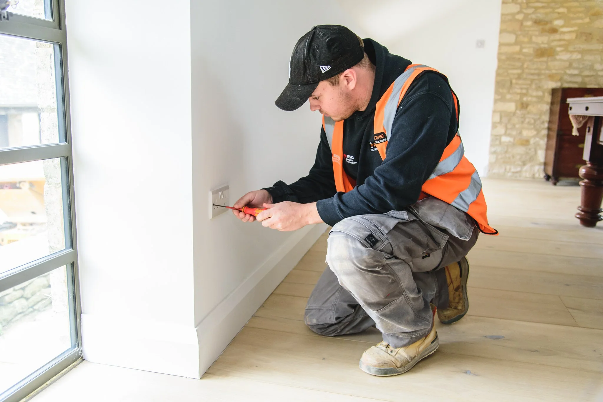 A worker kneeling on a wooden floor, wearing a black cap, orange safety vest, gray cargo pants, and beige work boots, inspecting or working on an electrical outlet near a glass window with a screwdriver.