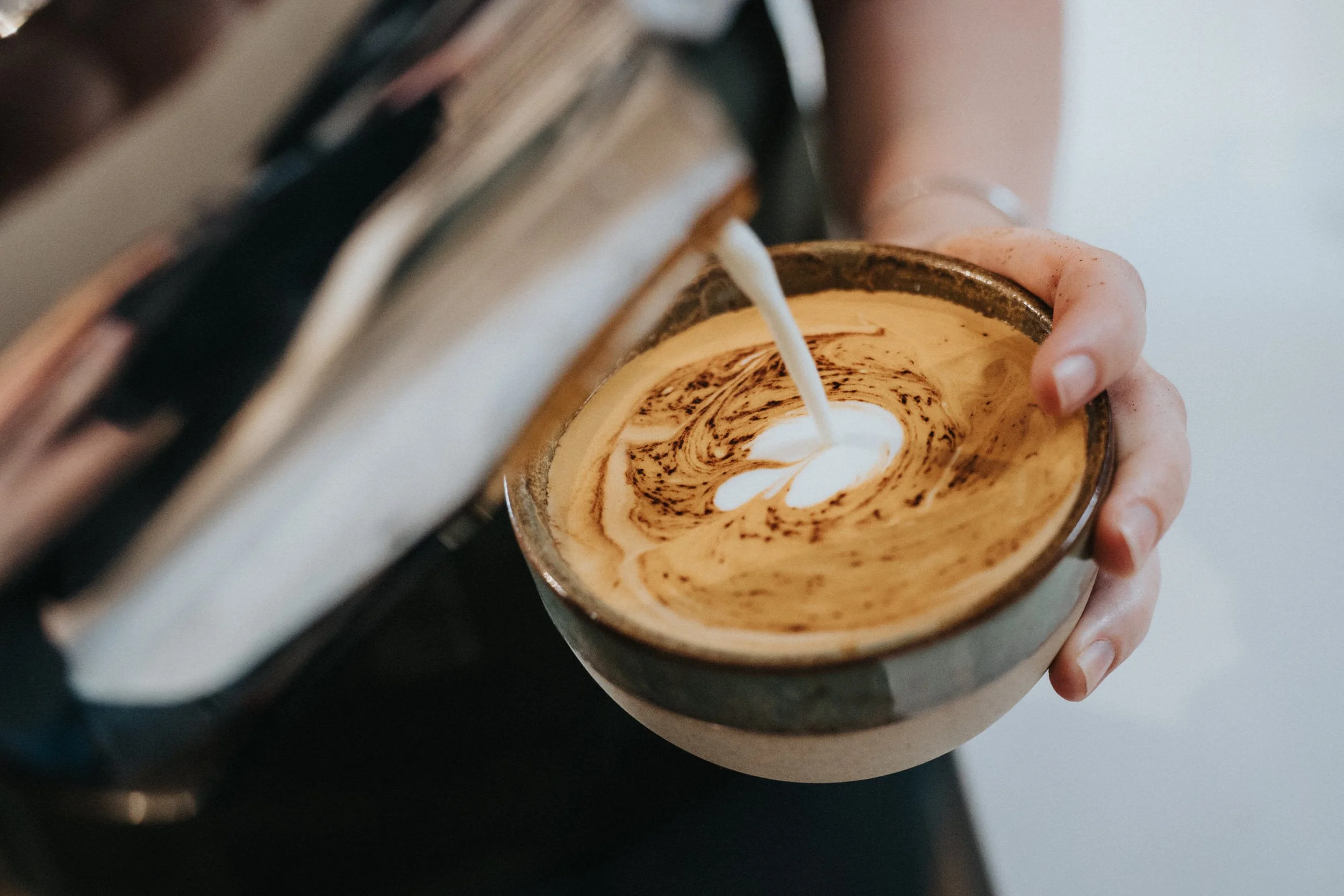 Person pouring steamed milk into a cup of espresso to make a latte, creating a swirl pattern on top.