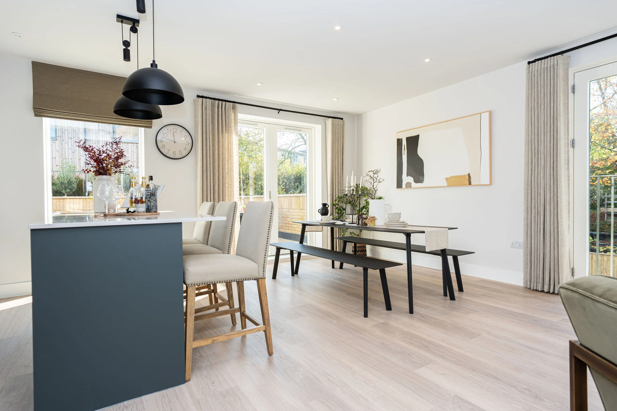 Modern dining room with a black and white abstract painting on the wall, a long table with benches, a bar area with cream chairs, large windows with curtains, and a sliding door leading outside, decorated with plants and minimalist decor.