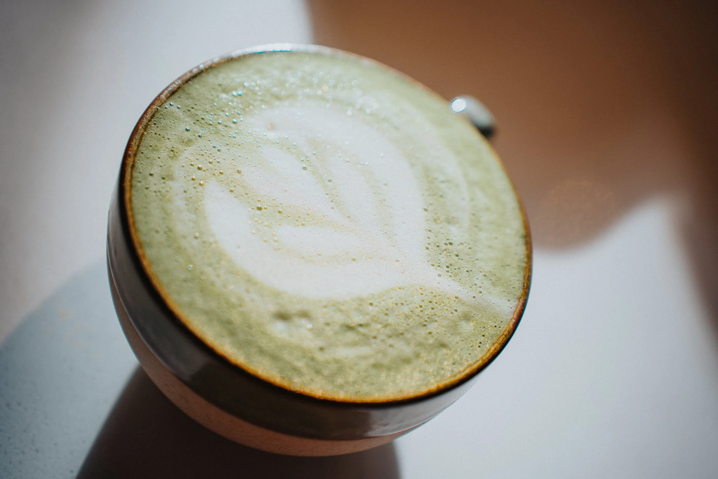 Close-up of a latte with foam art in a dark ceramic mug, with a shadow cast on a white surface.