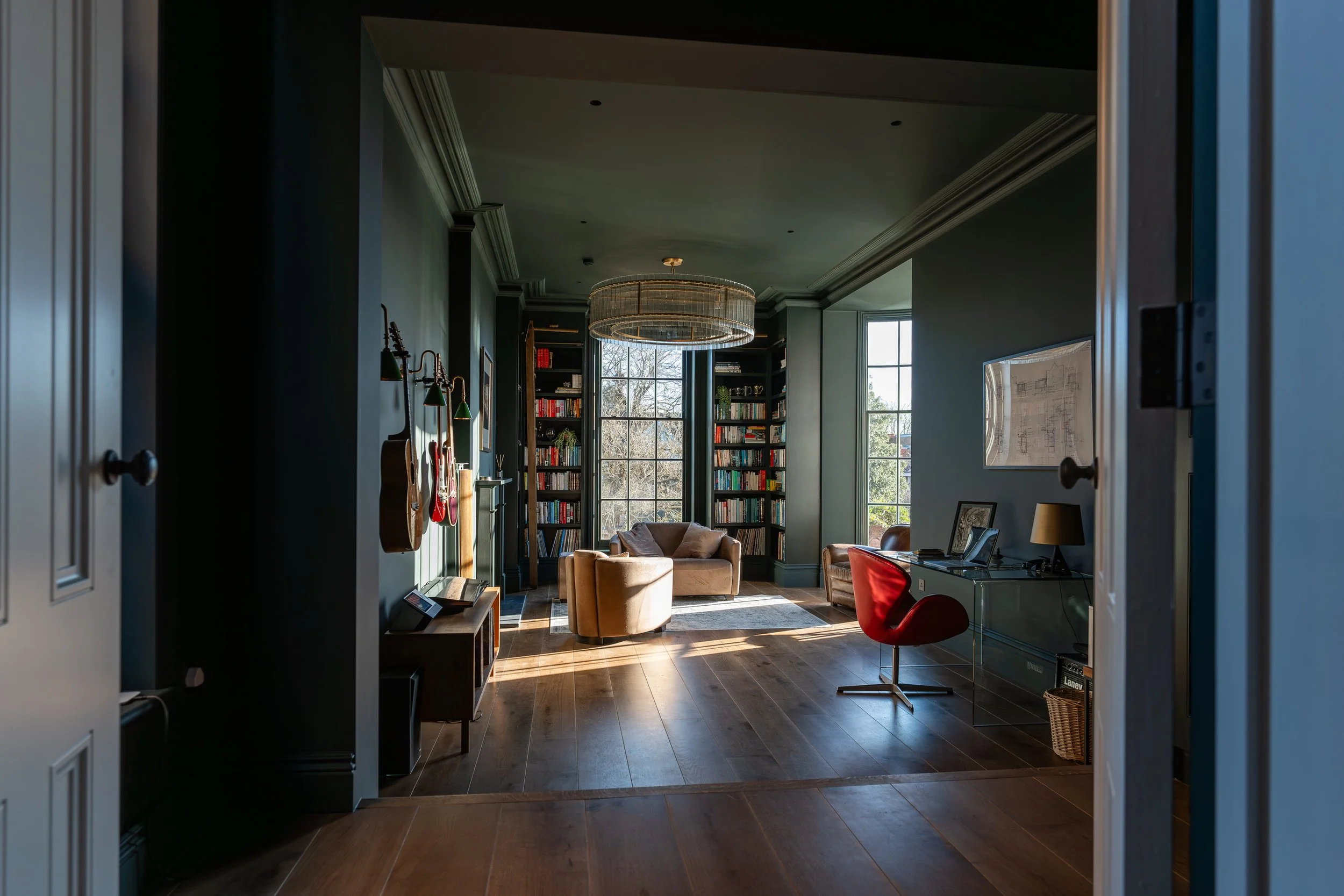 A cozy home office or library with a large window, bookshelves, a brown armchair, a Red chair, and a chandelier in a well-lit room.