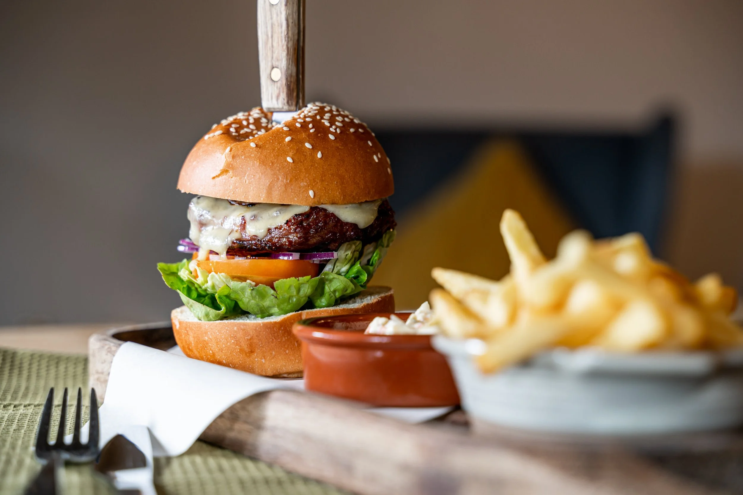 A cheeseburger with lettuce, tomato, onion, and melted cheese, served with French fries in a bowl, on a wooden board with a fork and knife.