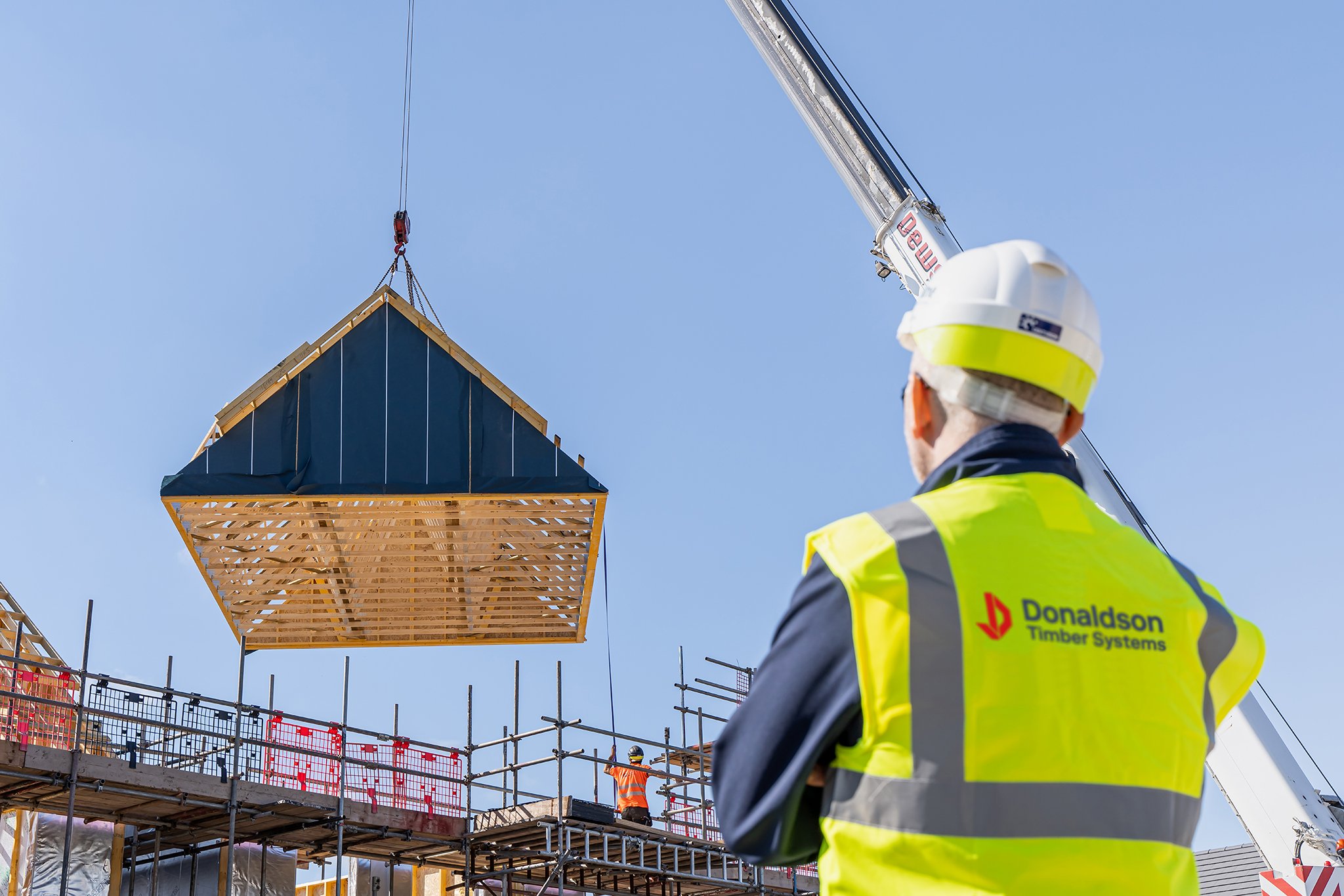 Construction worker in a yellow safety vest and white hard hat watching a crane lift a large wooden structure into place on a construction site with scaffolding and another worker in orange safety gear in the background.