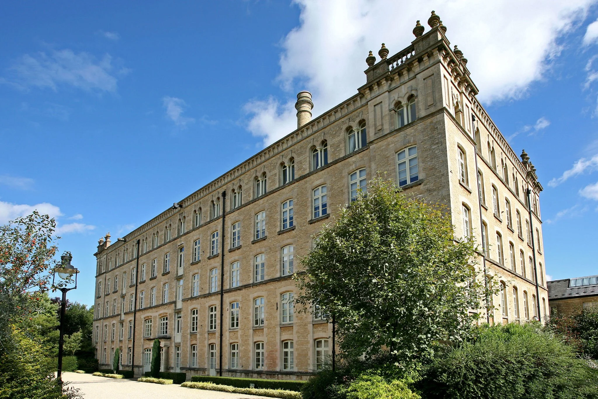 A historic stone building with multiple windows, surrounded by greenery and trees, under a partly cloudy blue sky.