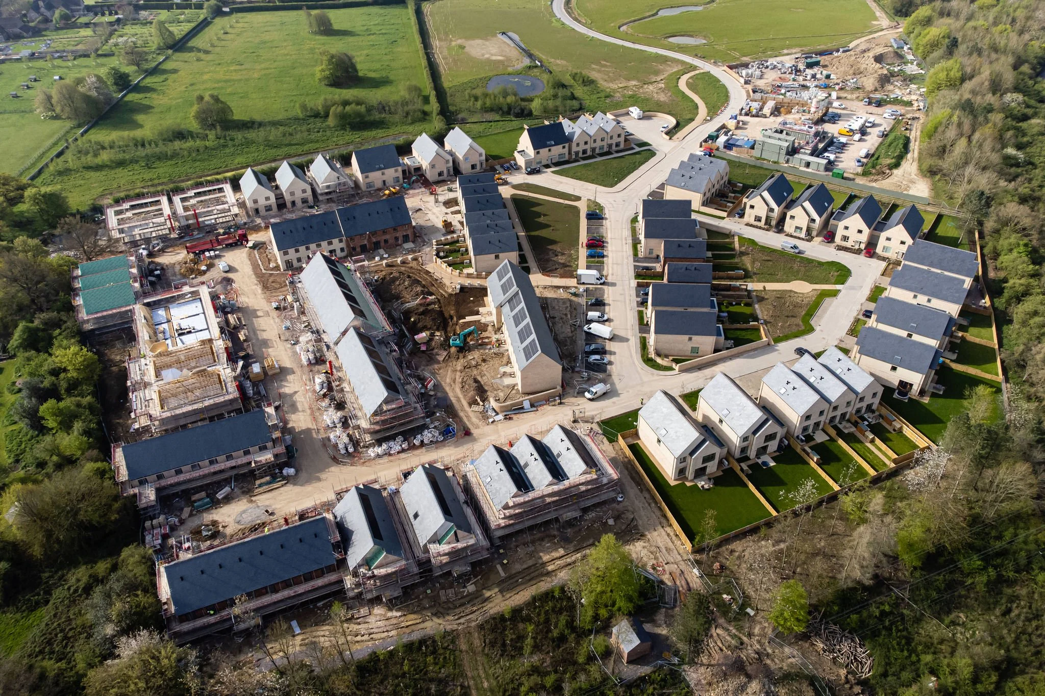 Aerial view of a residential housing development under construction with completed houses and ongoing building work surrounded by green fields and trees.