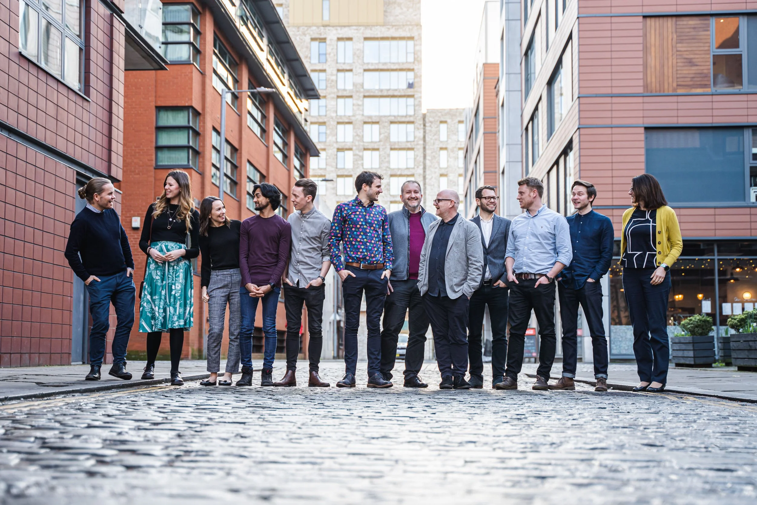A diverse group of people having their Team Shot image taken, dressed in business casual attire, standing in a city street with modern buildings in the background, engaging in conversation and smiling.