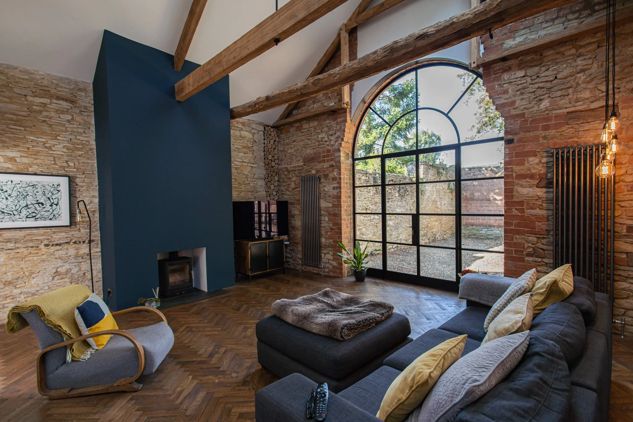 Living room with exposed brick walls, wooden beams, large arched Crittall window, and modern gray sofa with yellow and white pillows.