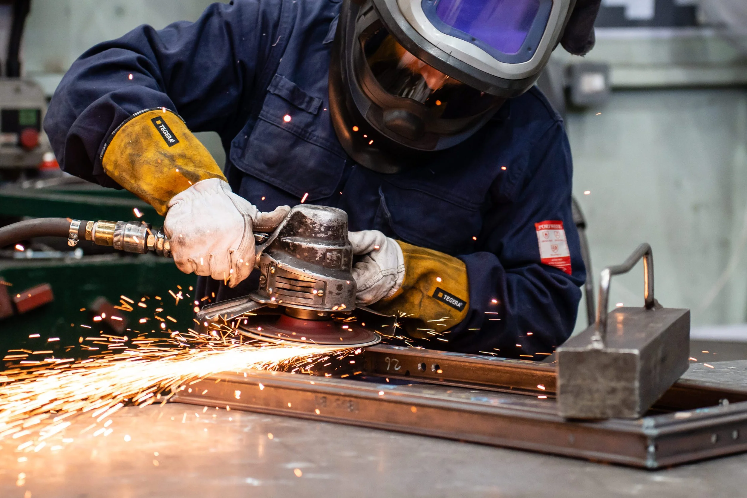 A person wearing protective gear and gloves using an angle grinder on metal Crittall frame, causing sparks to fly in an industrial workshop.
