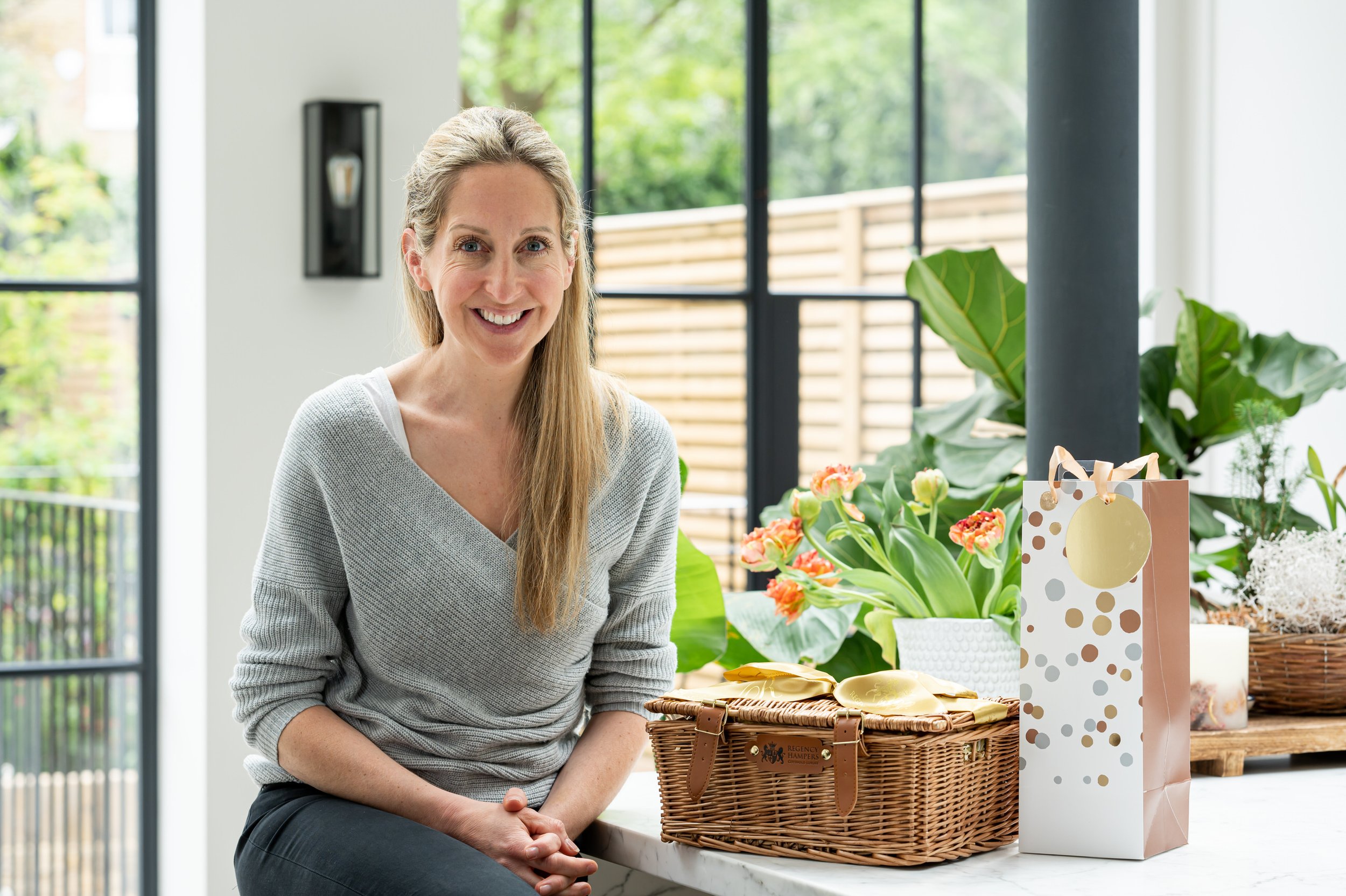 A smiling woman posing for a public relations photograph, receiving a prize with long blonde hair, in a gray sweater, sitting at a table in a bright, modern room with large windows. Flower plants and gift bags are on the table.