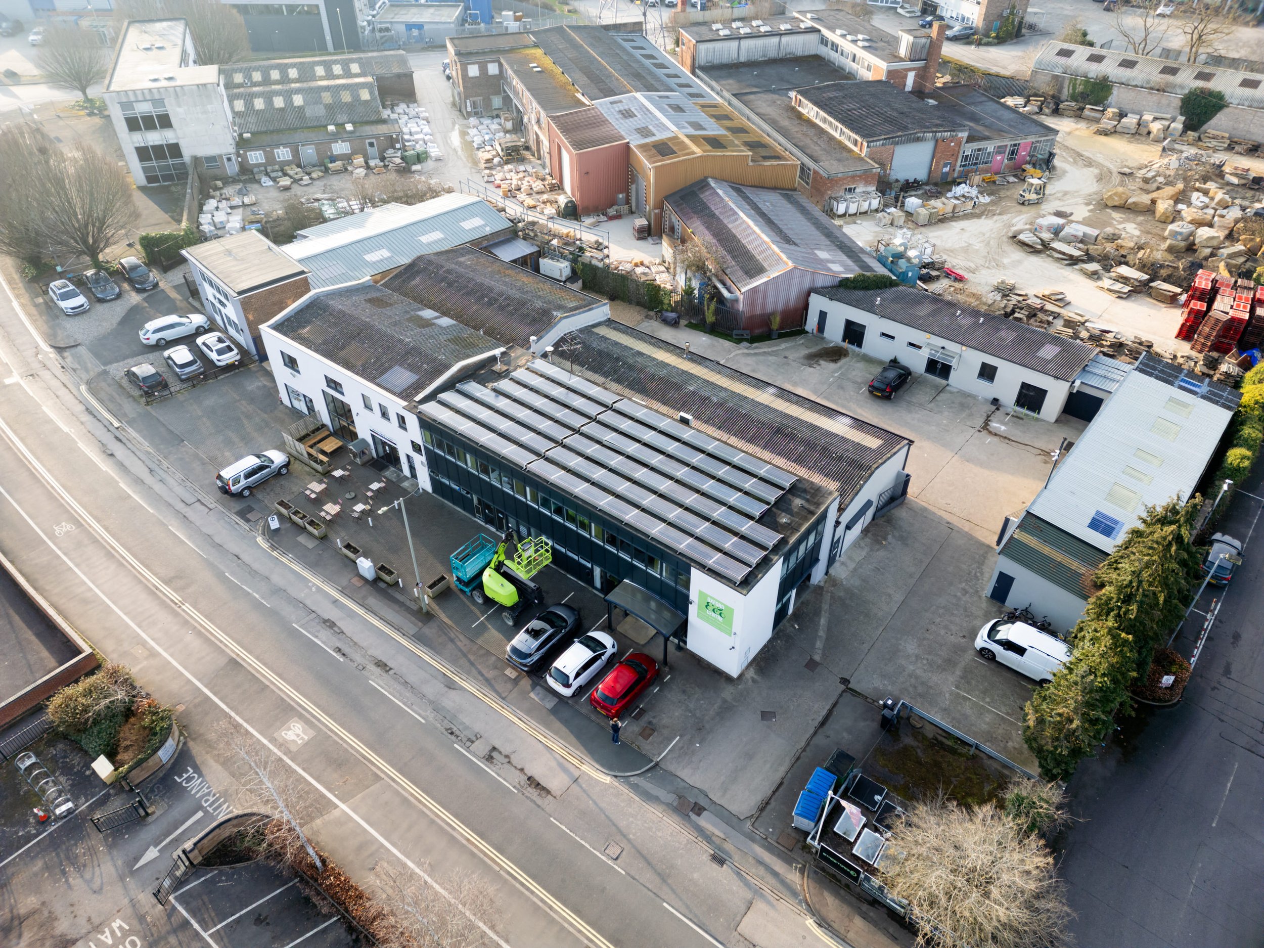 Aerial view of an industrial area with multiple commercial buildings, parking lots, some cars, and construction materials, located alongside a street with bike lanes.