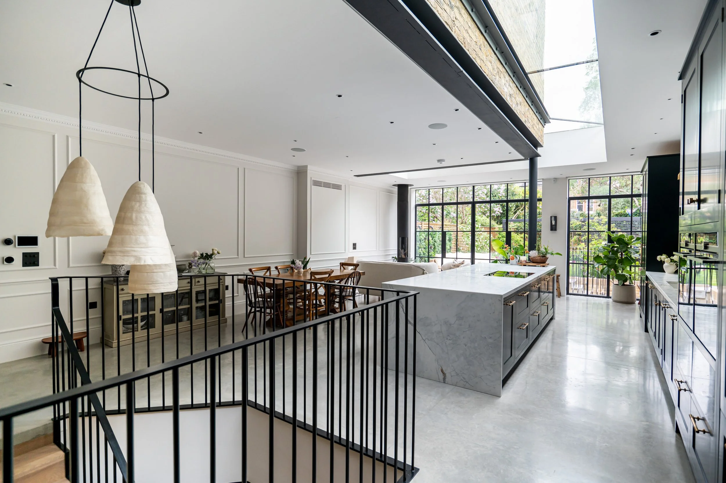 Modern open-concept kitchen and dining area with large Crittall windows, marble island, black cabinets, and wooden dining table with chairs.
